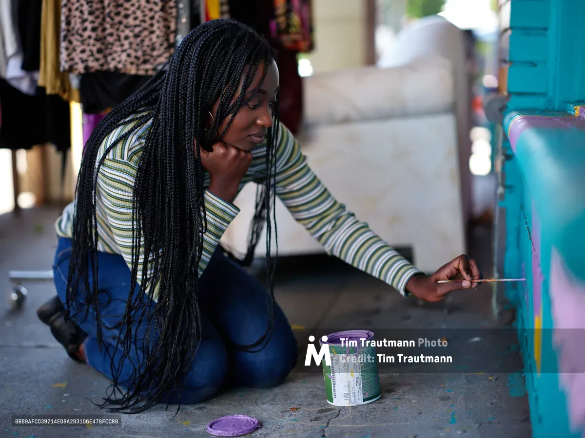 Naomi Likayi sits cross-legged on the concrete sidewalk, delicately applying her signature to a vibrant turquoise mural panel. Her long braids cascade over a striped shirt as afternoon light filters through the urban marketplace atmosphere of Portland's Alberta Arts District. An open paint can with purple pigment sits beside her, evidence of the creative work just completed. The scene captures an intimate moment of artistic completion against the backdrop of Northeast Portland's culturally rich community.