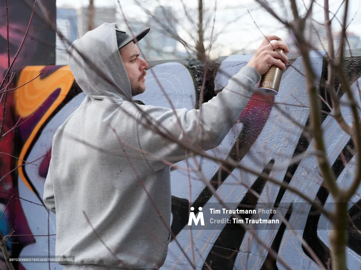 Travis Fields, known as Campographic, concentrates intently on his mural work at the Clay Creative Building in southeast Portland's Hosford-Abernethy neighborhood. Clad in a gray hoodie and snapback cap, the artist grips his spray paint can with focused determination against the concrete wall. Bare winter branches frame the scene in the foreground, while vibrant orange and purple graffiti elements provide a colorful backdrop to this moment of urban artistic creation.