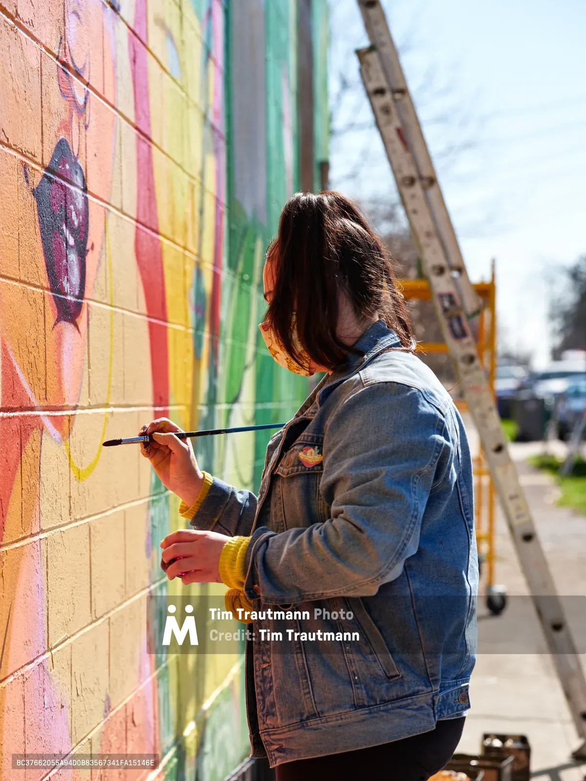 Multidisciplinary artist Villatose applies precise brushstrokes to her vibrant mural 'Jardín Cubana' depicting three female figures along Northeast Alberta Street in Portland's celebrated arts district. Dressed in a worn denim jacket and mustard sweater, the artist works with focused concentration against the colorful brick wall, her dark hair catching the afternoon light. The scene captures the intimate process of public art creation, with scaffolding and urban infrastructure framing this moment of creative expression in one of Portland's most dynamic cultural neighborhoods.