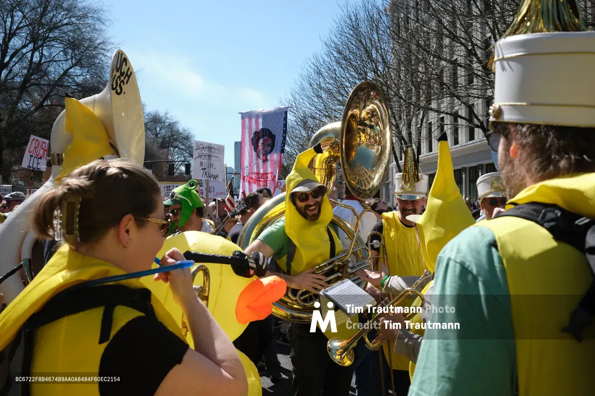 A spirited brass ensemble clad in vibrant yellow ponchos performs amid a political demonstration in Portland's historic Old Town district. The tubist at center, wearing sunglasses and a broad grin, anchors the musical protest as fellow musicians brandish their instruments like banners of resistance. Behind them, protest signs punctuate the crowd while bare winter trees frame the scene against a crisp blue sky, creating a striking tableau of civic engagement through music.