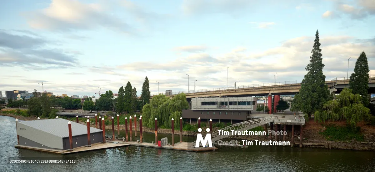 Portland Fire & Rescue Station 21 sits anchored on the Willamette River beneath the gentle light of an overcast evening sky. The modern floating firehouse, distinguished by its corrugated metal roof and red support pilings, connects to shore via a steel gangway, creating a unique waterfront emergency response facility. Towering Lombardy poplars frame the scene while the Hawthorne Bridge spans the background, establishing the station's strategic location in Portland's Central Eastside industrial district.