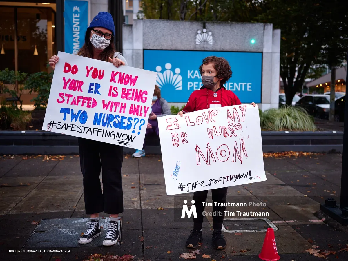 A woman in a cobalt blue knit beanie and face mask holds a handwritten protest sign questioning emergency room staffing levels, while a young child beside her displays a heartfelt message of support for their nurse mother. The pair stands on autumn-scattered sidewalks outside Kaiser Permanente's towering headquarters in Portland's Lloyd District, their white poster boards catching the soft urban light against the healthcare giant's signature blue corporate signage. The intimate scale of their grassroots demonstration contrasts powerfully with the institutional architecture looming behind them.