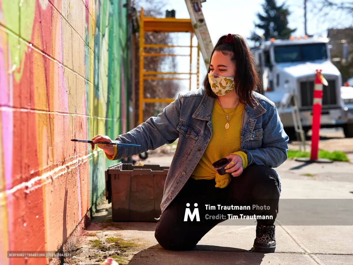 Multidisciplinary artist Vilatose kneels on the sidewalk, paintbrush in hand, adding vibrant details to 'Jardín Cubana,' a colorful mural depicting three female figures along a brick wall in Portland's Alberta Arts District. Bathed in warm afternoon sunlight, she wears a yellow knit sweater beneath a denim jacket and a patterned face mask, her dark hair pulled back as she focuses intently on her work. Behind her, construction equipment and urban infrastructure frame this moment of artistic creation in the heart of Northeast Portland's creative corridor.