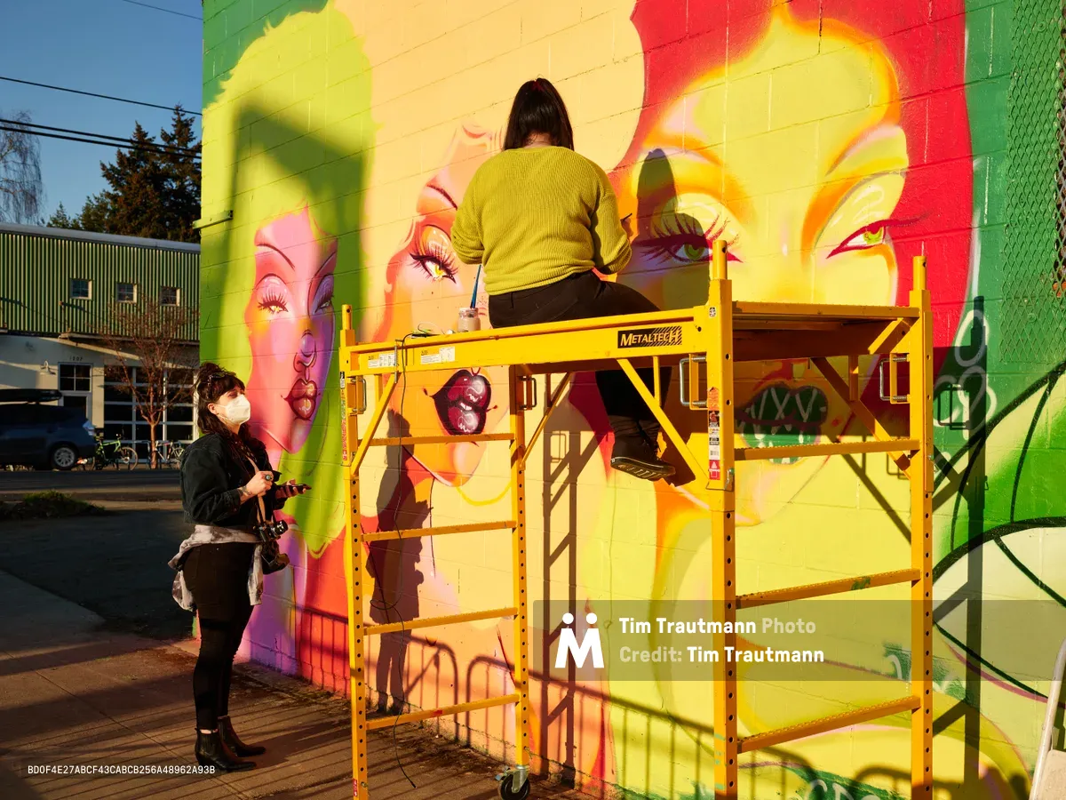 Artist Villatose works from a yellow scissor lift to paint 'Jardín Cubana,' a vibrant mural depicting three female figures on a brick wall along Northeast Alberta Street in Portland's Alberta Arts District. A masked collaborator documents the process from below, capturing the creative energy of this urban art installation. The mural features bold yellows, pinks, and greens that transform the building's facade, while late afternoon light creates dramatic shadows across the industrial equipment and colorful artwork.