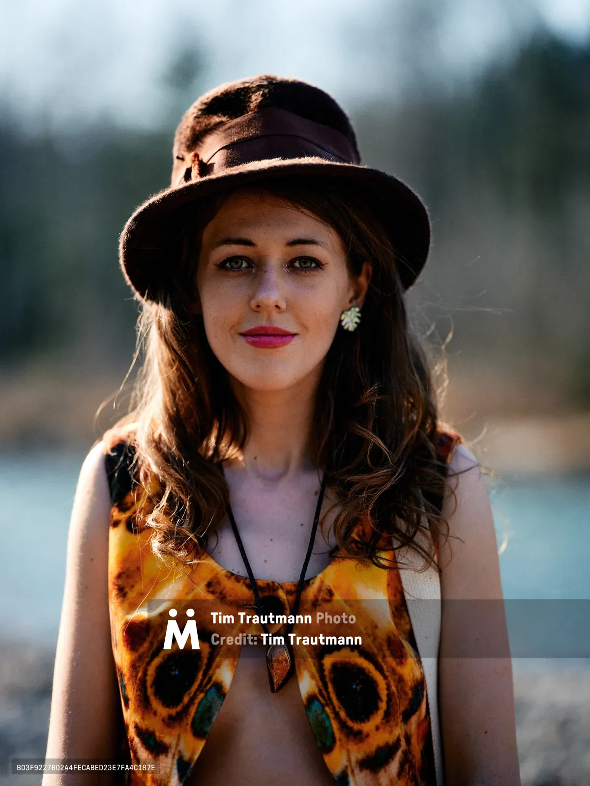 A young woman with flowing brunette hair poses confidently along the Floodplain Trail riverbed in Multnomah County, Oregon, her striking butterfly-wing printed top catching the warm afternoon light. Her wide-brimmed felt hat casts gentle shadows across her face while bold pink lipstick and delicate pearl earrings add vintage elegance to her bohemian style. The soft bokeh background of Pacific Northwest riverbed landscape creates an dreamy, ethereal atmosphere that highlights her direct gaze and natural beauty.