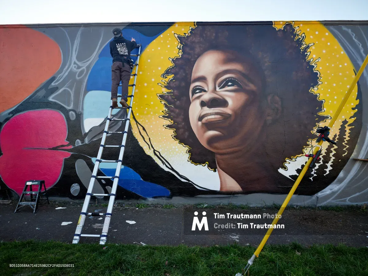 Artist Travis Fields, known as campographic, stands atop an aluminum ladder while painting a vibrant mural of poet Amanda Gorman on a wall in Northeast Portland's Alberta district. The luminous portrait depicts Gorman with her natural hair flowing against a golden celestial backdrop dotted with stars, her gentle smile radiating hope and determination. The surrounding composition features bold geometric shapes in coral, magenta, and azure blue, creating a dynamic frame around the central figure. Paint supplies and equipment scattered on the weathered pavement below speak to the ongoing creative process transforming this urban canvas.