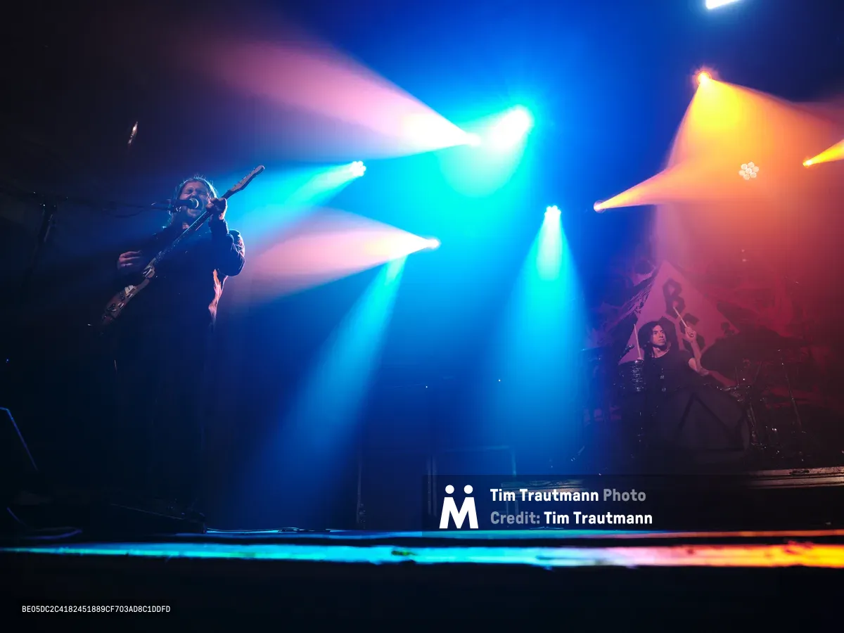 Russell Marsden and Julian Dorio of Band of Skulls perform during their 10 Year Anniversary Tour at Webster Hall in Manhattan's East Village. Intense cyan and amber stage lights slice through the atmospheric darkness, creating dramatic beams that illuminate the musicians in silhouette. The legendary venue's intimate setting amplifies the raw energy as the duo delivers their signature sound beneath the venue's historic ceiling, with stage smoke adding ethereal texture to the lighting design.