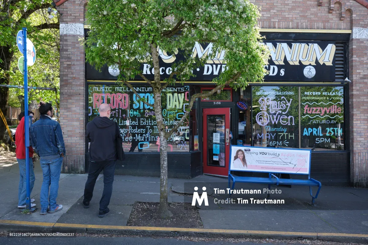 Two men pause on the sidewalk outside Music Millennium, a beloved Portland record store on East Burnside Street, their casual stance suggesting the unhurried rhythm of neighborhood browsing. Hand-painted window advertisements for Record Store Day and upcoming performances create a vibrant collage against the brick storefront, while dappled sunlight filters through spring foliage overhead. The scene captures the enduring appeal of independent music retail in Portland's eclectic Kerns neighborhood, where vinyl culture thrives amid the city's creative community.