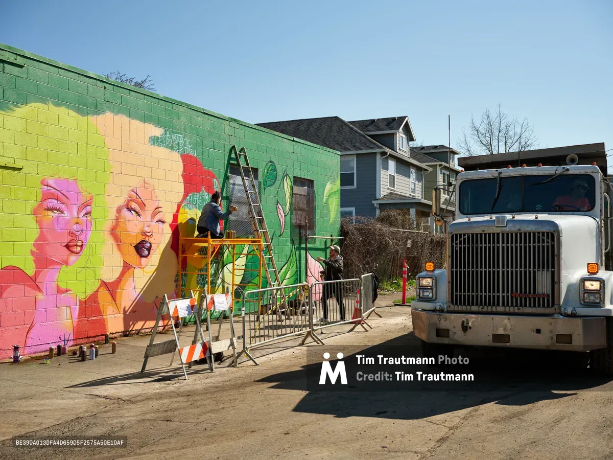 Multidisciplinary artist Villatose works from scaffolding to paint a vibrant pop art mural titled 'Jardín Cubana' featuring three stylized female figures on a green cinder block wall along Northeast Alberta Street in Portland's Alberta Arts District. The scene captures the intersection of urban art and community development, with residential houses visible in the background and construction equipment stationed nearby. Bright afternoon light illuminates the bold pink, yellow, and red palette of the emerging artwork, while safety barriers and a white commercial truck frame this moment of creative transformation in the gentrifying neighborhood.