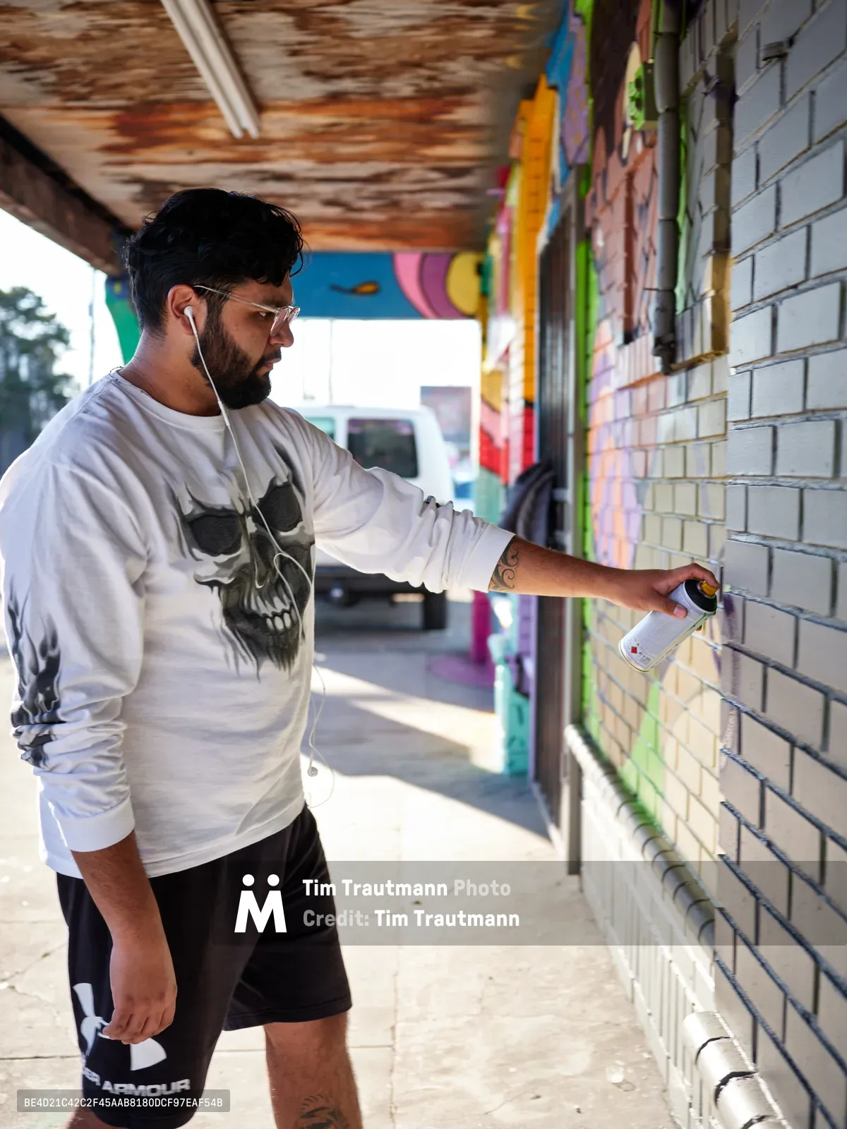 Muralist Heysus concentrates intently as he applies spray paint to a weathered brick wall beneath a covered walkway in Portland's Alberta district. The bearded artist, wearing a skull-adorned white sweatshirt and earbuds, works methodically against the textured urban canvas while vibrant existing murals create a kaleidoscope of color in the background. Dappled sunlight filters through the overhead structure, casting dramatic shadows that emphasize the gritty authenticity of this Northeast Portland street art scene.