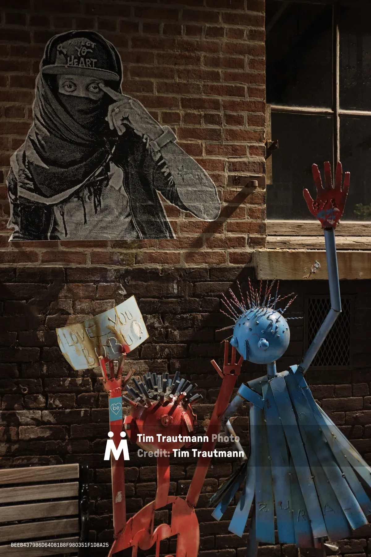 A provocative street art tableau unfolds against weathered brick in Brooklyn's Williamsburg neighborhood, where anonymous rebellion meets whimsical protest. A masked figure rendered in stark black and white wheat paste dominates the wall, bearing the cryptic message 'NY HEART,' while below, two vibrant sculptural figures—one crimson, one azure—raise defiant hands skyward in a gesture of solidarity. The afternoon light catches the textured surfaces and peeling paint, creating a dialogue between the ephemeral nature of street art and the permanent urban landscape.