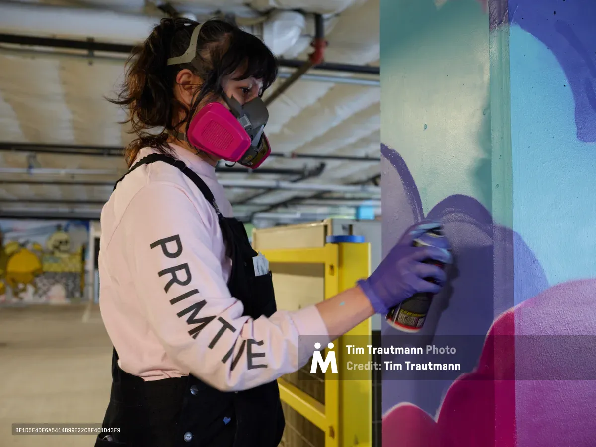 A focused muralist wearing a pink respirator mask and "PRIMITIVE" sweatshirt applies purple spray paint to a vibrant wall mural in the converted parking garage of Clay Creative in Southeast Portland's Hosford-Abernethy neighborhood. The industrial workspace, with its exposed ceiling infrastructure and yellow storage units, provides an authentic urban canvas for the emerging artwork's bold blues, purples, and magentas.