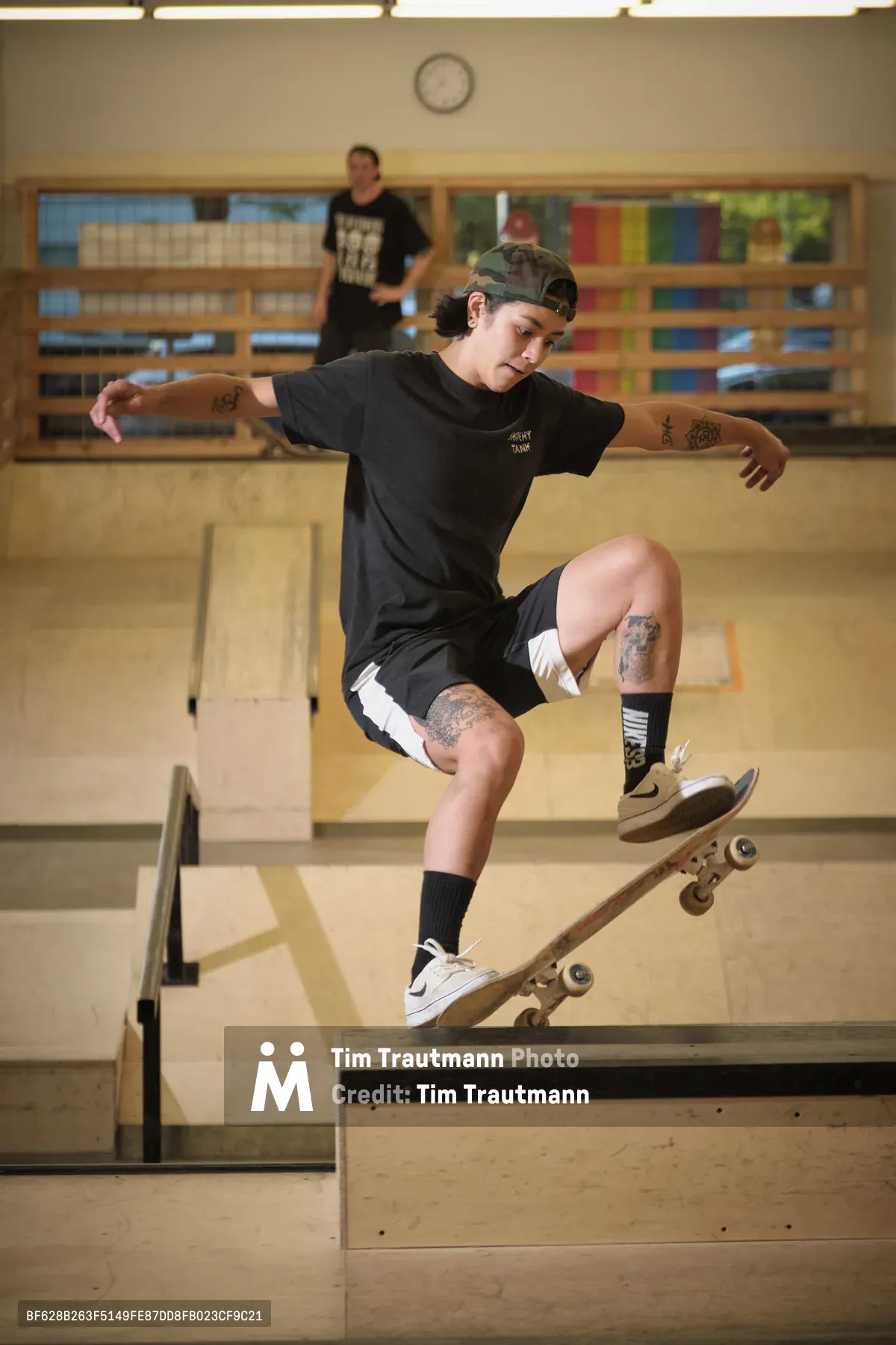 Pro skateboarder Christiana Means executes a precise frontside carve across the curved concrete of Stronger Skatepark in Milwaukie, Oregon. Her tattooed arms extended for balance, she navigates the bowl's transition with fluid control while fluorescent lights cast an even glow across the indoor facility. The geometric lines of the skate park's wooden ramps create a backdrop of angles and shadows, with another skater visible in the distance.