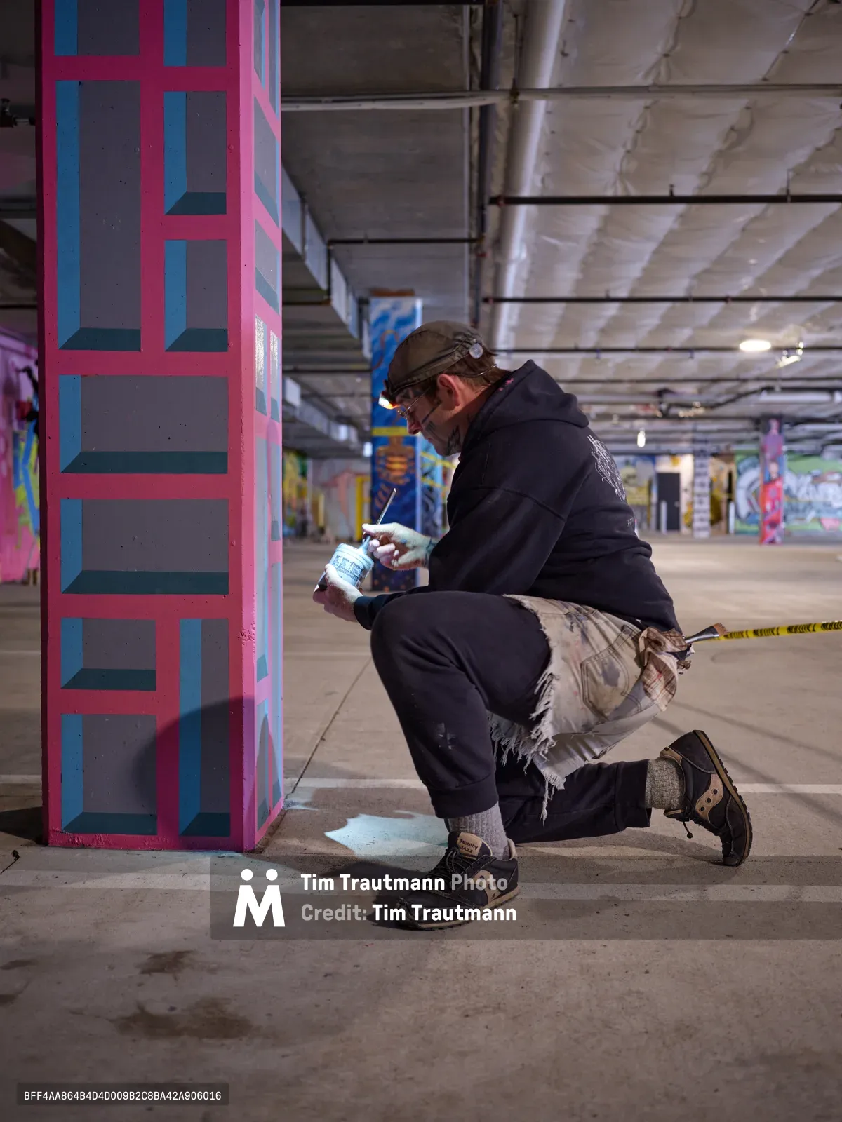 Portland artist and bike activist Rabbitt kneels in focused concentration within the Clay Creative building's parking garage, spray painting his signature geometric patterns onto a concrete column. The harsh fluorescent lighting casts dramatic shadows across the industrial space, illuminating vibrant pink and blue geometric forms that transform the utilitarian concrete into urban canvas. His weathered work clothes and protective gear speak to the physical dedication required for large-scale mural creation in Portland's creative underground.