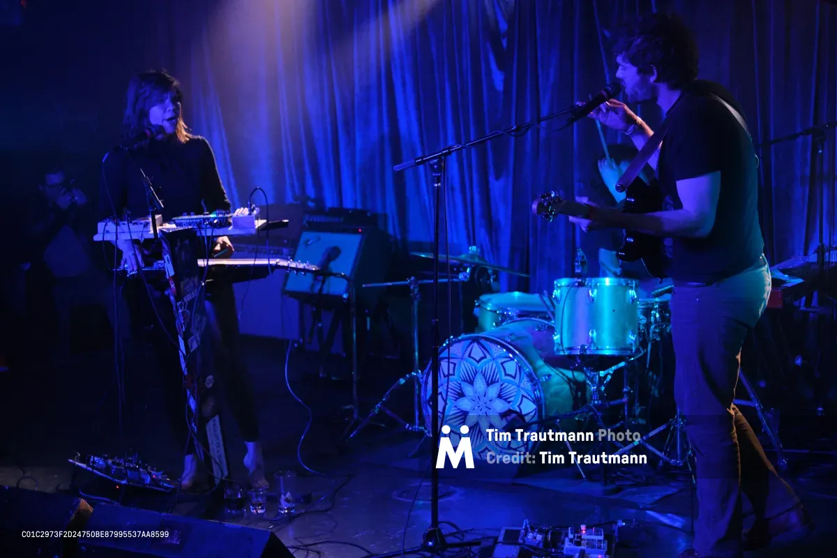 Experimental band Sun Blood Stories, originally from Boise, Idaho and now based in Portland, performs in the intimate confines of Dante's venue during Rx Fest, bathed in deep cobalt stage lighting that transforms the space into an ethereal sonic cathedral. The keyboardist, silhouetted against flowing curtains, commands an array of synthesizers while the drummer maintains rhythm behind a kit adorned with geometric mandala artwork. Cables snake across the stage floor as purple and blue washes create dramatic shadows, capturing the mesmerizing live sound that has been likened to modern-day opera.