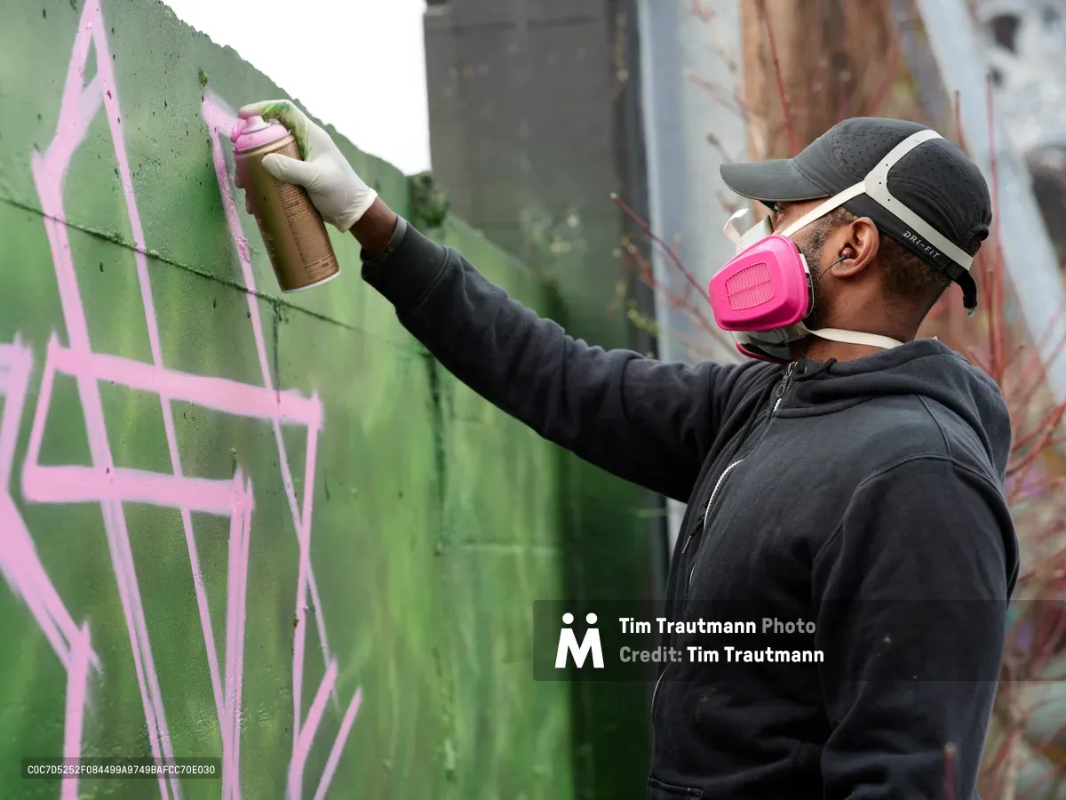 A dedicated muralist wearing a bright pink respirator mask and gray hoodie applies fresh pink paint to a vibrant green wall in the parking lot of Clay Creative in Portland's Hosford-Abernethy neighborhood. The artist's focused concentration is evident as he carefully guides the spray can across the concrete surface, adding geometric lines to what appears to be an emerging street art piece. The industrial setting and protective gear speak to the serious craft of contemporary urban art creation.