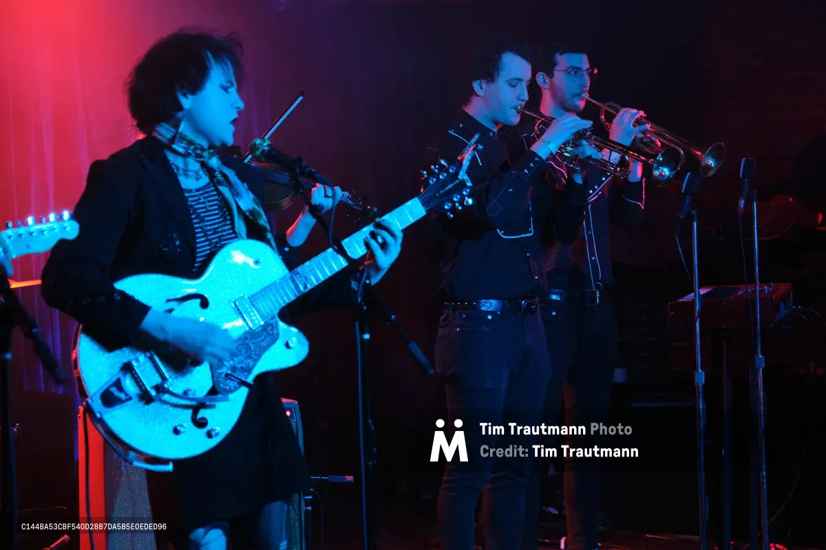 Charlotte McCaslin of Roselit Bone commands the stage with her hollow-body electric guitar, bathed in saturated cyan and crimson stage lighting at Dante's in Portland. The intimate venue's moody atmosphere envelops the trio as brass players behind her contribute to the sonic landscape. The dramatic color temperature creates an otherworldly glow against the dark venue backdrop, capturing the raw energy of Portland's underground music scene.