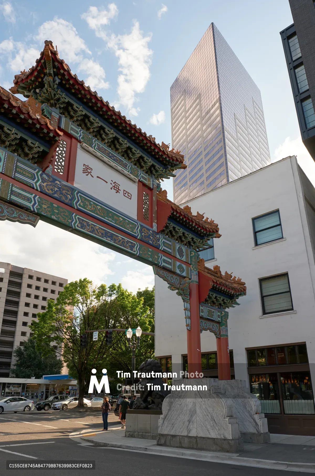 A magnificent Chinese paifang gateway stands sentinel at the entrance to Portland's Chinatown, its coral-red pillars and ornate jade-green brackets creating a striking counterpoint to the gleaming glass tower rising behind it. The traditional architecture, adorned with intricate decorative elements and Chinese characters, bridges centuries as pedestrians move beneath its ceremonial arch. Soft afternoon light bathes the scene, highlighting the cultural juxtaposition between ancient craftsmanship and contemporary urban development in this historic Pacific Northwest neighborhood.