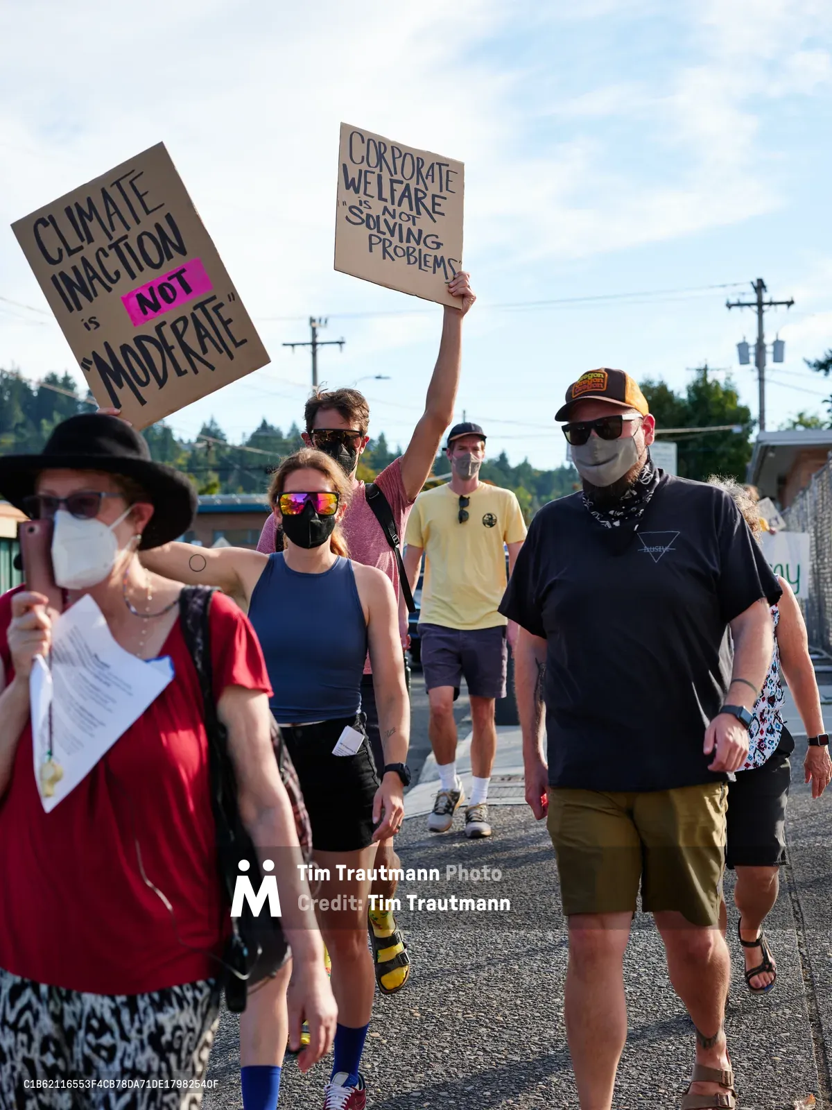Masked protesters stride purposefully down a sun-dappled street in Oregon City, Oregon, their handmade signs demanding climate action and critiquing corporate welfare. The diverse group of activists moves with quiet determination beneath a partly cloudy sky, their colorful summer clothing contrasting against the stark black lettering of their protest signs. Power lines stretch overhead while residential buildings frame this grassroots demonstration for environmental justice.