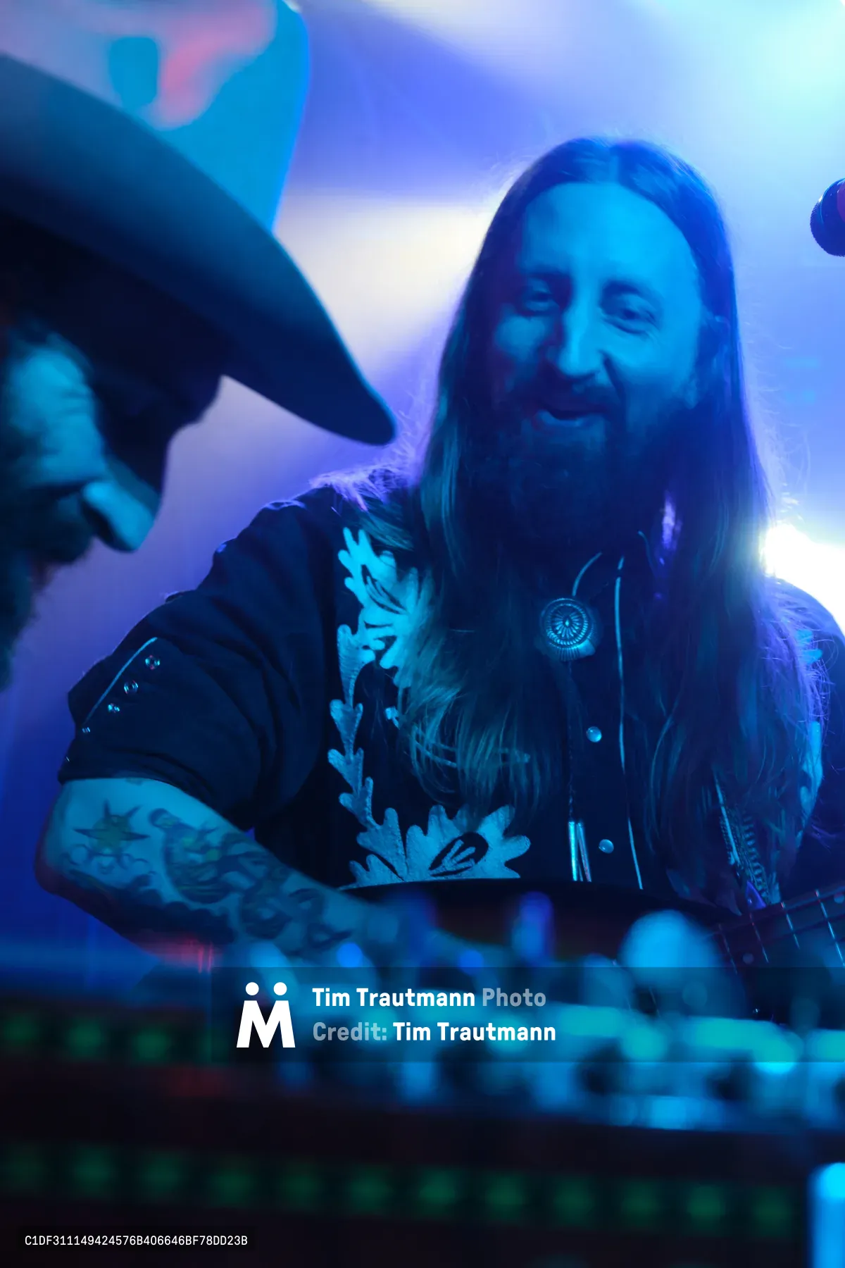 A bearded musician with flowing hair grins widely while performing behind an electronic mixing console, bathed in vibrant blue and purple stage lighting. The intimate venue atmosphere pulses with energy as the artist, part of electronic duo Roselit Bone, manipulates controls during their RX Fest 2019 performance. Tattoos visible on his arms catch the atmospheric lighting while the background dissolves into a dreamy wash of concert illumination.