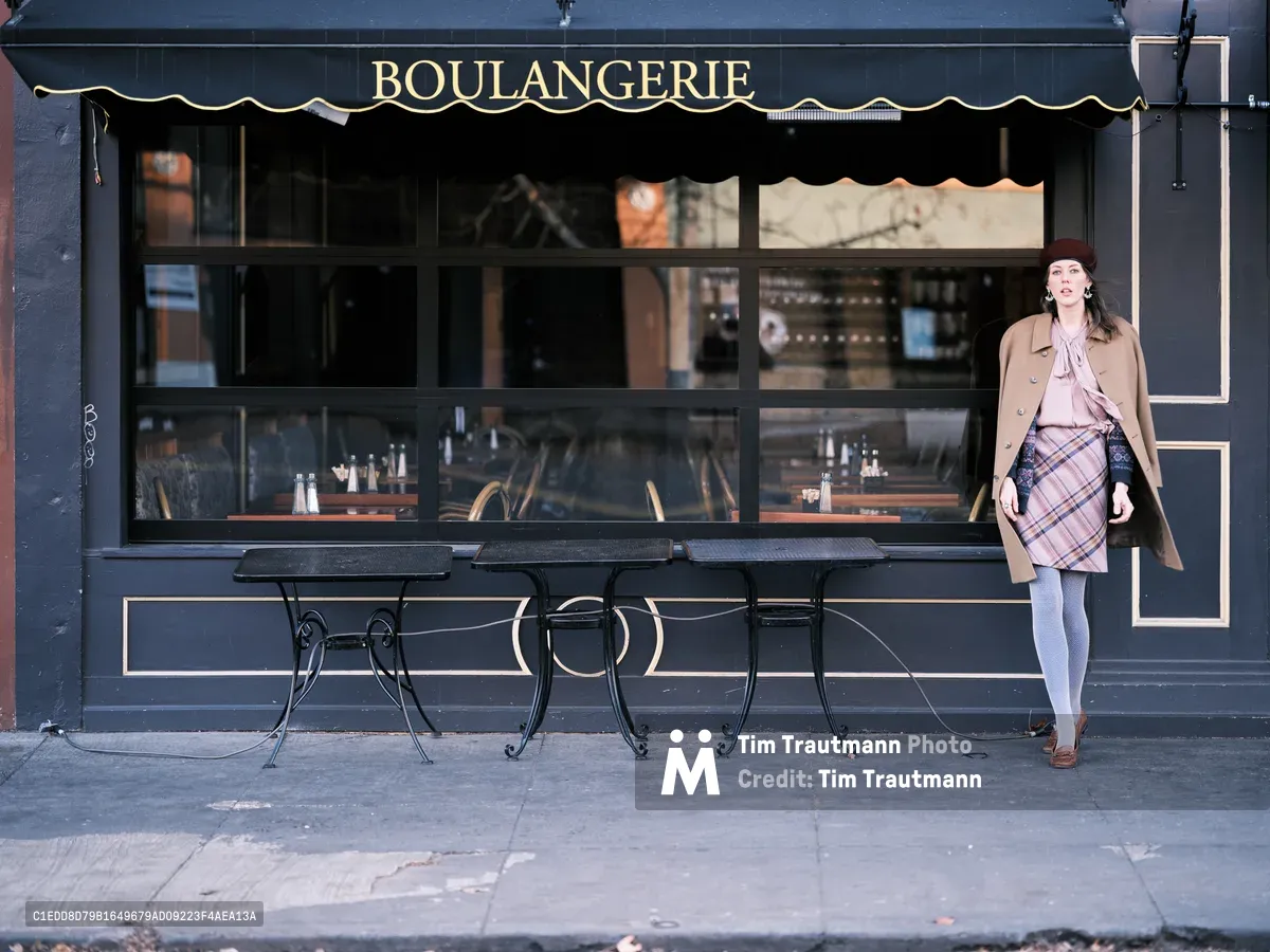 A stylish woman in a burgundy beret, camel coat, and plaid skirt stands beside a charming French bakery on Northeast Alberta Street in Portland, Oregon. The scene captures the intersection of European café culture and Pacific Northwest urban life, with wrought iron bistro tables positioned beneath the boulangerie's classic black awning. Soft natural light illuminates the storefront's large windows, revealing glimpses of the bakery's interior while creating an intimate street-side moment that speaks to Portland's vibrant neighborhood dining culture.