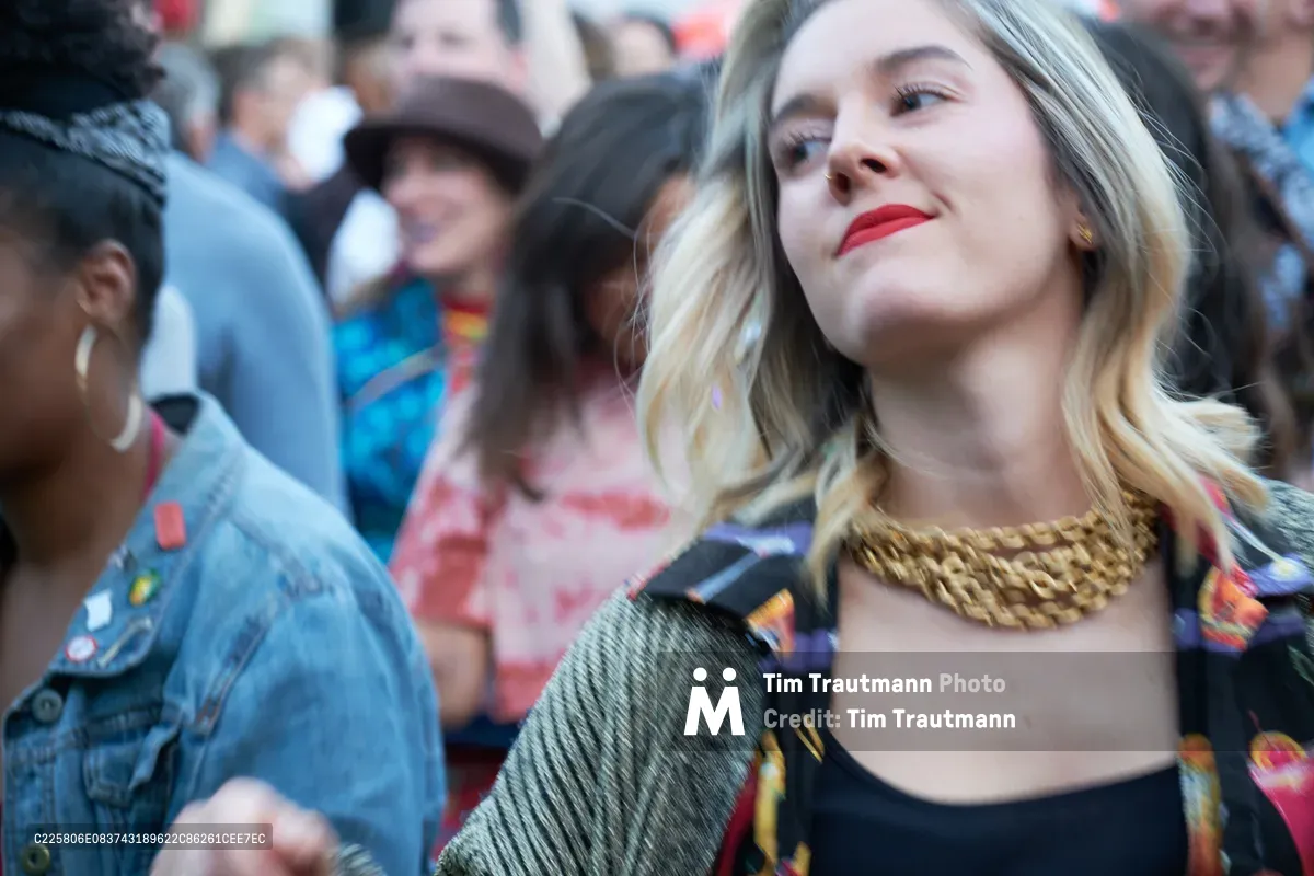 A young blonde woman with crimson lips and a statement gold chain necklace gazes upward with serene satisfaction, captured in the warm glow of late afternoon light at White Owl Social Club's summer day party. The shallow depth of field blurs the denim-clad crowd behind her into soft bokeh, creating an intimate portrait against the bustling Portland social scene. Her metallic chain catches the golden light while partygoers fade into impressionistic shapes of blue and coral hues.