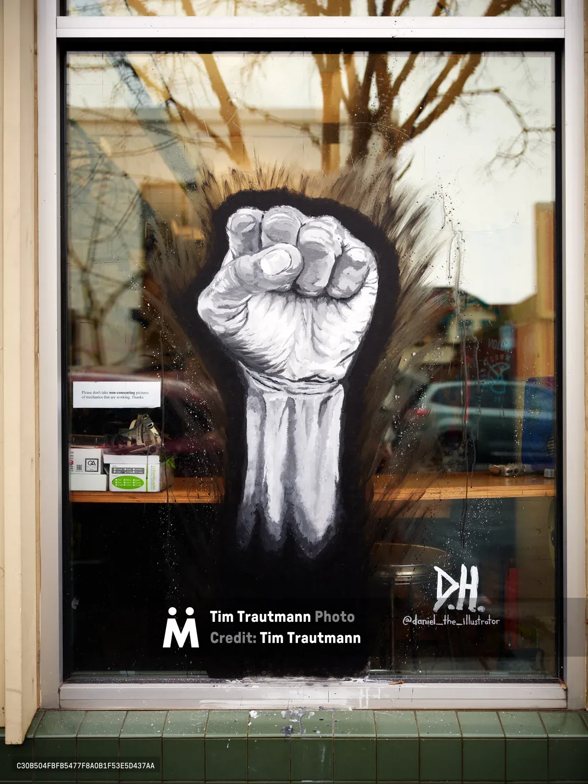 A powerful monochromatic raised fist emerges from an explosive dark burst, painted across the storefront window of the Community Cycling Center on Northeast Alberta Street in Portland's Alberta District. The dramatic black and white artwork dominates the glass surface, with the clenched fist rendered in striking detail against radiating brushstrokes that suggest both energy and resistance. Reflections of bare winter trees and the surrounding streetscape create layered transparency over the bold political imagery, while the artist's signature "DH" appears in the lower right corner.