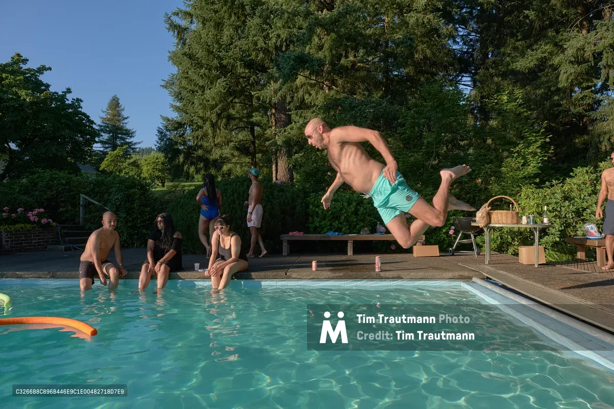 A shirtless man in mint green swim shorts launches into a crystal-clear swimming pool mid-leap, his body captured in dynamic motion against the backdrop of towering evergreens. Friends gather poolside in this intimate backyard gathering at Orchard Vibration in Newberg, Oregon, where golden afternoon light filters through the verdant canopy. The scene radiates carefree summer joy, with pool toys floating in the turquoise water and picnic essentials scattered on nearby tables. The lush Pacific Northwest landscape frames this moment of pure recreational bliss.