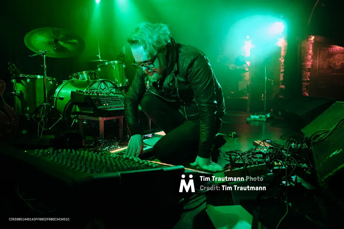 Electronic musician Eli Savage, performing as Ellipsism, hunches intently over his mixing console at RXFest 2019, bathed in ethereal green stage lighting that transforms Dante's intimate venue into a sonic laboratory. His silver hair catches the emerald glow as he manipulates analog controls, surrounded by a constellation of synthesizers, drum machines, and electronic equipment that sprawl across the darkened stage. The atmospheric lighting creates dramatic shadows and highlights, emphasizing the concentrated focus of the artist as he sculpts soundscapes in Portland's legendary underground music venue.