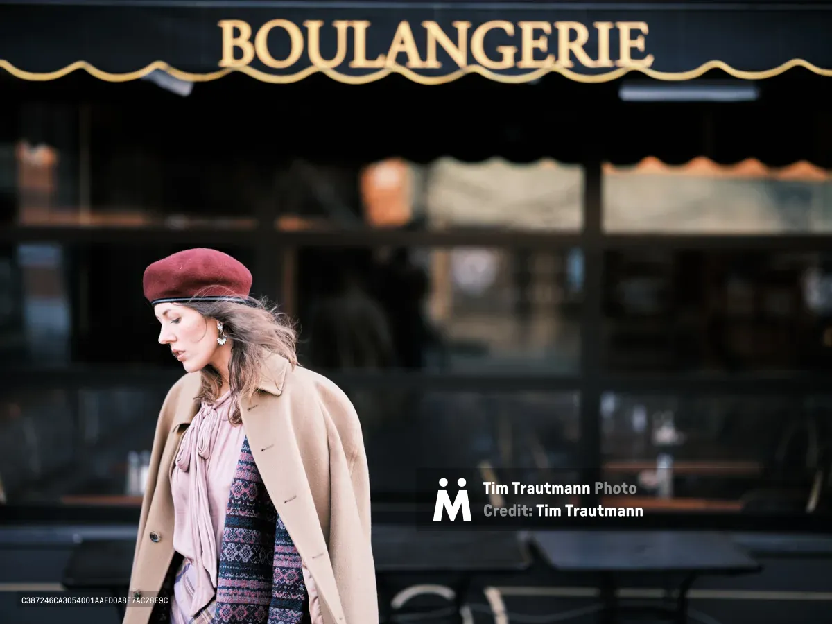 A woman in a burgundy beret and camel coat strolls past a classic French boulangerie storefront on Northeast Alberta Street in Portland's vibrant arts district. The golden afternoon light illuminates her contemplative profile as she moves through this slice of European charm transplanted to the Pacific Northwest. Her layered autumn ensemble of neutral tones contrasts beautifully against the deep forest green awning and traditional bakery signage.
