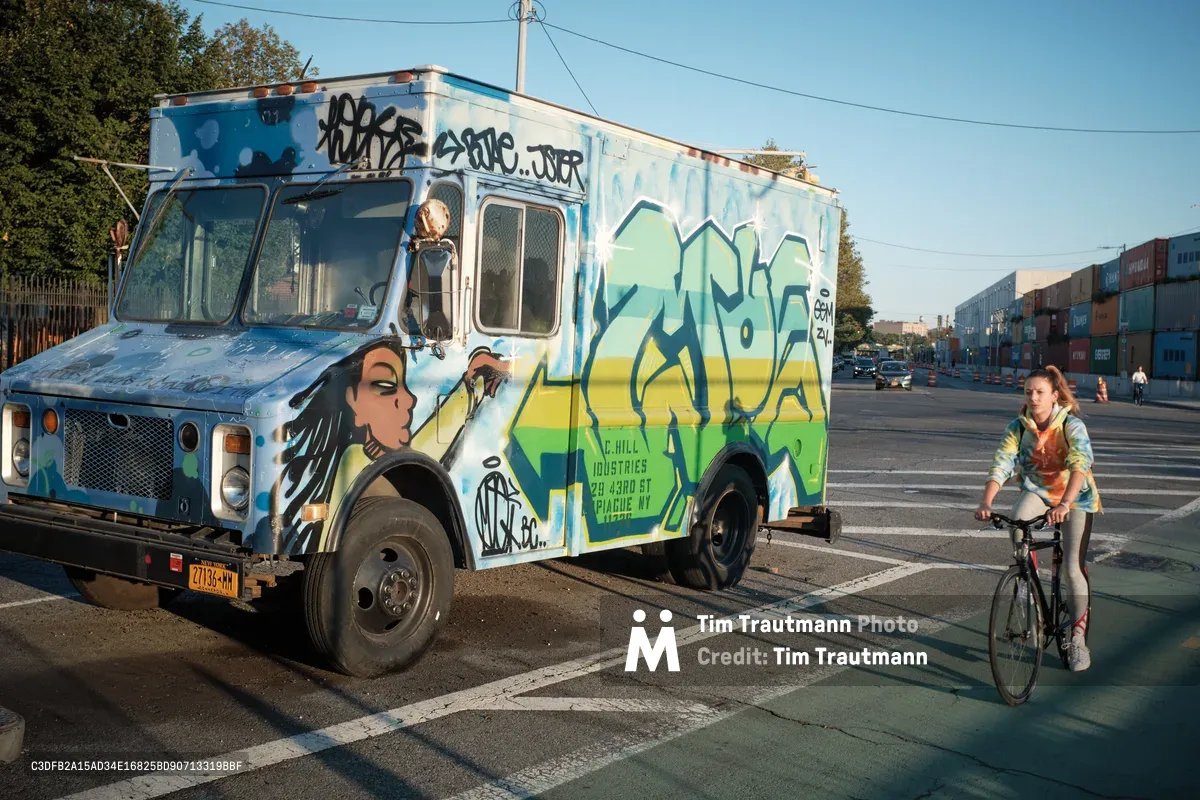 Golden hour light bathes a vibrant graffiti-covered food truck parked along Kent Avenue in Brooklyn's Navy Yard district, its weathered white surface transformed by bold blues, yellows, and greens depicting stylized figures and tags. A young woman on a bicycle pauses beside the mobile canvas, her figure casting long shadows across the asphalt as industrial shipping containers stack in the soft-focused background. The scene captures the raw creative energy of Brooklyn's waterfront, where street art culture intersects with everyday urban life.