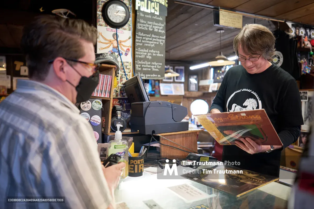 Inside the iconic Music Millennium record store on East Burnside in Portland's Kerns neighborhood during Record Store Day 2022, a bespectacled shopkeeper in a black t-shirt carefully examines a vinyl album while a masked customer waits at the counter. The intimate scene unfolds beneath warm pendant lighting, surrounded by towering shelves of records and memorabilia. A vintage wall clock and handwritten store policy sign create an authentic atmosphere of analog music culture, while the glass counter displays rare finds below.