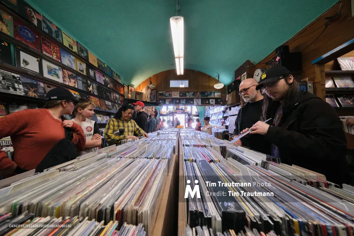 Music enthusiasts of varying ages browse through densely packed vinyl record bins at Music Millennium on East Burnside Street in Portland, Oregon. The intimate record shop features mint green walls lined floor-to-ceiling with album covers, creating a colorful mosaic of music history. Fluorescent lighting illuminates the focused faces of collectors as they flip through thousands of records, their bodies leaning into the ritual of vinyl discovery in this beloved neighborhood institution.