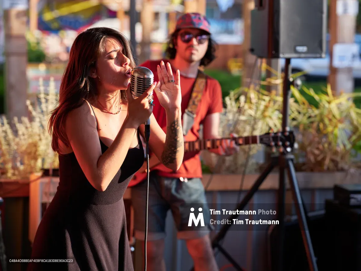 Aubrey Gordon commands the vintage chrome microphone during an intimate outdoor performance at Portland Mercado in Southeast Portland. The warm, golden light of late afternoon bathes the scene as the vocalist delivers her set with passionate intensity, her tattooed arm extended in expressive gesture. Behind her, a guitarist in a red flannel shirt and patterned cap provides accompaniment, while the venue's colorful backdrop of food carts and urban architecture creates an authentic Portland atmosphere.
