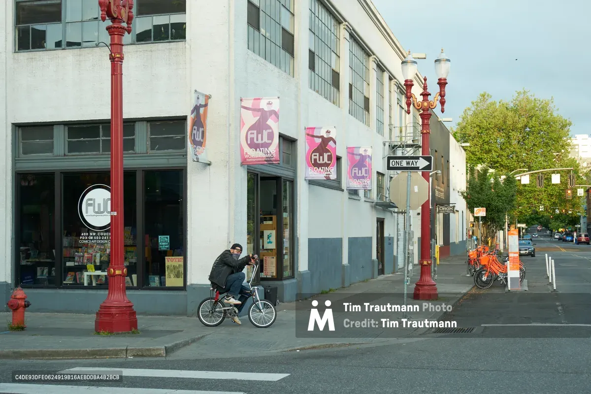 A masked cyclist navigates the quiet streets of Portland's Chinatown-Japantown Historic District during early morning hours. The scene captures the intersection of modern urban life and historic preservation, with ornate red Victorian-era street lamps standing sentinel against a backdrop of converted industrial buildings now housing creative businesses. Pink promotional banners for a local establishment flutter in the morning breeze, while the distant silhouette of downtown Portland creates depth in the soft, hazy light.