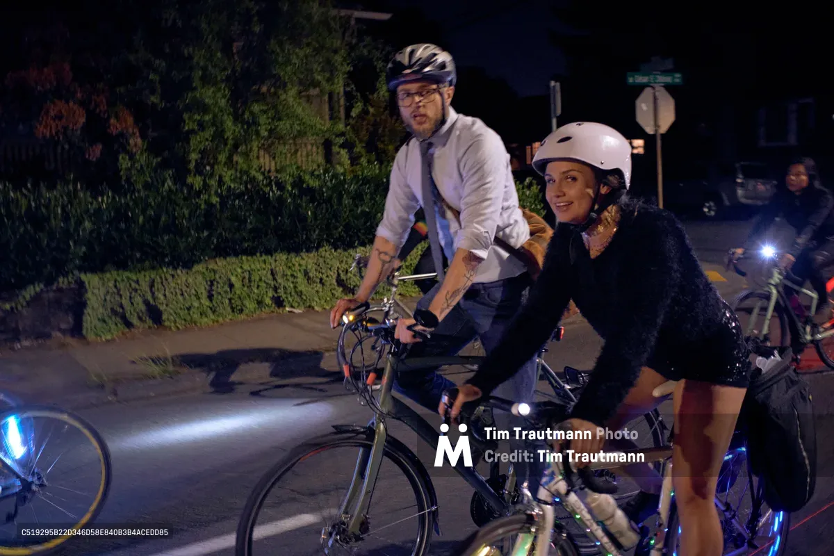 Two helmeted cyclists pause during Portland's Drop Out Prom Ride, part of the city's annual Pedalpalooza festival in June 2019. The man in a white dress shirt and tie contrasts with the woman in dark attire as they navigate the nighttime streets of Southeast César E. Chávez Boulevard. LED bicycle lights pierce the darkness while lush greenery creates a natural backdrop, capturing the playful intersection of formal wear and urban cycling culture during this unconventional prom celebration.