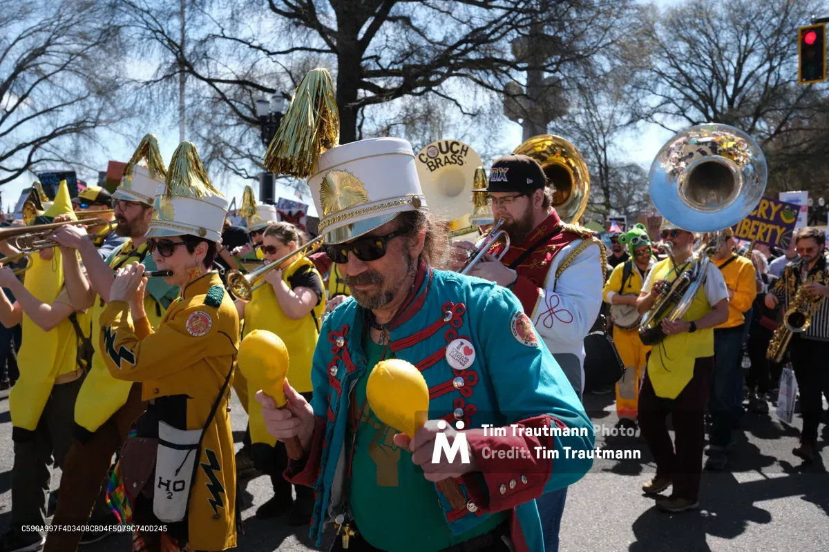 The Unpresidented Brass Band leads a spirited anti-monarchy demonstration through Portland's Skidmore Historic District, their yellow uniforms and gleaming instruments creating a defiant symphony against bare winter trees. A bearded musician in turquoise and red holds twin maracas while his white ceremonial hat catches the crisp daylight, surrounded by fellow protesters wielding tubas and trumpets in organized rebellion. The scene pulses with grassroots energy as demonstrators carrying protest signs merge with the musical procession along Southwest Ankeny Street.