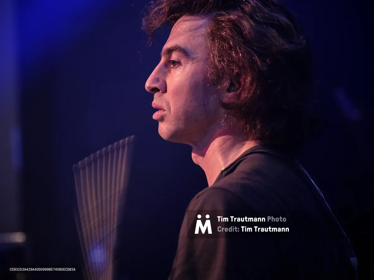 Julian Dorio, drummer for Band of Skulls, captured in profile during the band's 10 Year Anniversary Tour at Webster Hall in Manhattan. Theatrical blue and purple stage lighting sculpts his concentrated expression as he gazes into the distance, his tousled hair catching the ambient glow. The intimate venue's atmosphere envelops the scene in deep shadows, with the suggestion of his drum kit visible in the soft focus background.