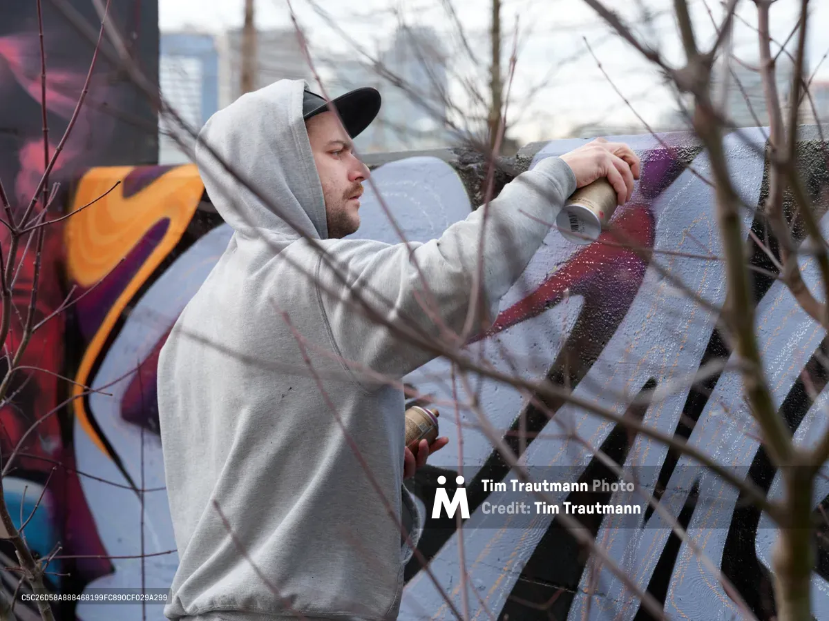 Travis Fields, known as Campographic, concentrates on his craft as he applies spray paint to a weathered concrete wall at the Clay Creative Building in southeast Portland's Hosford-Abernethy neighborhood. Bare winter branches create a natural frame in the foreground, while his grey hoodie and baseball cap provide stark contrast against the emerging purple and red hues of his urban artwork. The overcast Pacific Northwest light filters through the scene, capturing this moment of artistic creation in Portland's vibrant street art community.