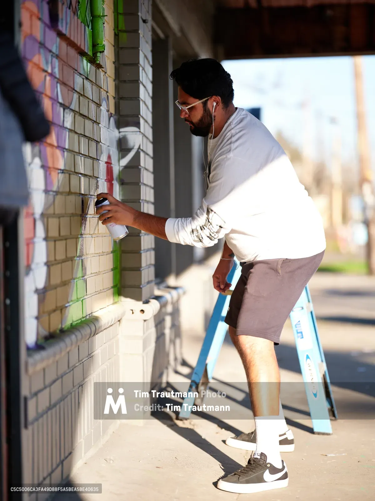 Artist Heysus concentrates intently as he applies spray paint to a cream-colored brick wall, working from a blue stepladder on a sun-drenched sidewalk in Northeast Portland. The warm afternoon light illuminates his focused expression and white long-sleeved shirt as he adds vibrant details to his emerging mural. Green tape marks his careful preparation work against the textured brick surface, while the soft-focus urban backdrop hints at the Alberta Arts District's creative energy.