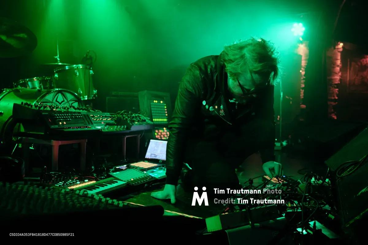 Electronic musician Eli Savage, performing as ellipsism, becomes fully absorbed in his craft at RXFest 2019, hunched over an intricate web of synthesizers and mixing equipment at Portland's intimate Dante's venue. Ethereal green stage lighting bathes the performance space, creating an otherworldly atmosphere that mirrors the experimental nature of his electronic compositions. His flowing hair catches the colored light as he manipulates controls with intense concentration, embodying the solitary focus required for live electronic music creation.