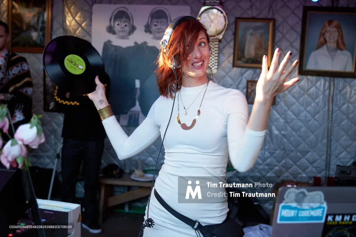 DJ Doctor Dakar commands the dance floor with infectious energy at White Owl Social Club, her auburn hair catching golden light as she holds a vinyl record aloft while gesturing enthusiastically to the crowd. The intimate Portland venue's eclectic wall art creates a bohemian backdrop of vintage portraits and quilted textures. Her white fitted top and layered jewelry embody the relaxed sophistication of Sunday afternoon party culture in the Pacific Northwest.