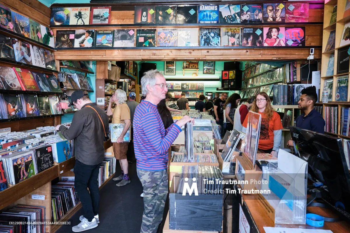 Inside the beloved Music Millennium on East Burnside, Portland's iconic record store pulses with afternoon energy as vinyl enthusiasts navigate densely packed aisles. A silver-haired collector in a blue striped shirt examines albums near towering wooden bins, while other customers browse the extensive collection that climbs floor-to-ceiling along warm wood shelving. The shop's mint green walls and eclectic atmosphere create an intimate sanctuary for music discovery.