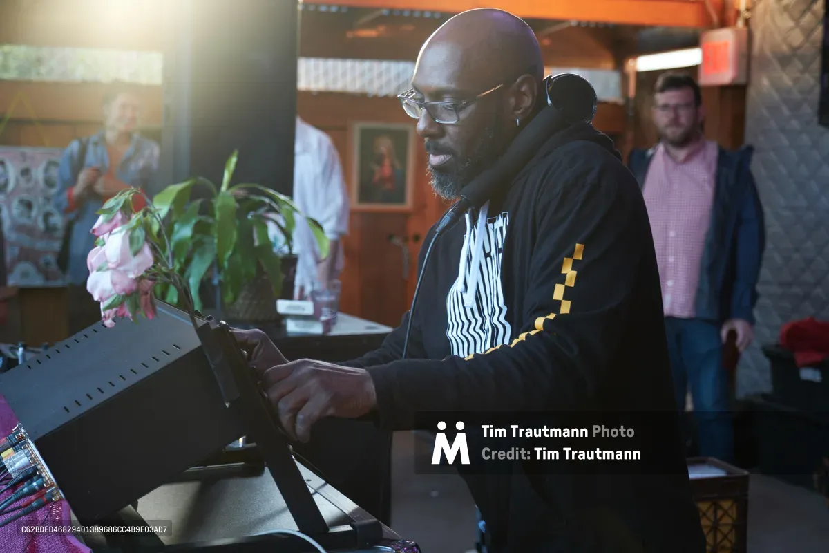DJ Cee White commands the turntables at White Owl Social Club during Your Sunday Best day party, his weathered hands working the equipment with practiced precision. Golden afternoon light streams through the Portland venue, illuminating his focused expression behind wire-rimmed glasses as patrons gather in the warm, intimate space. Fresh flowers accent the DJ booth while the eclectic club atmosphere buzzes with anticipation.