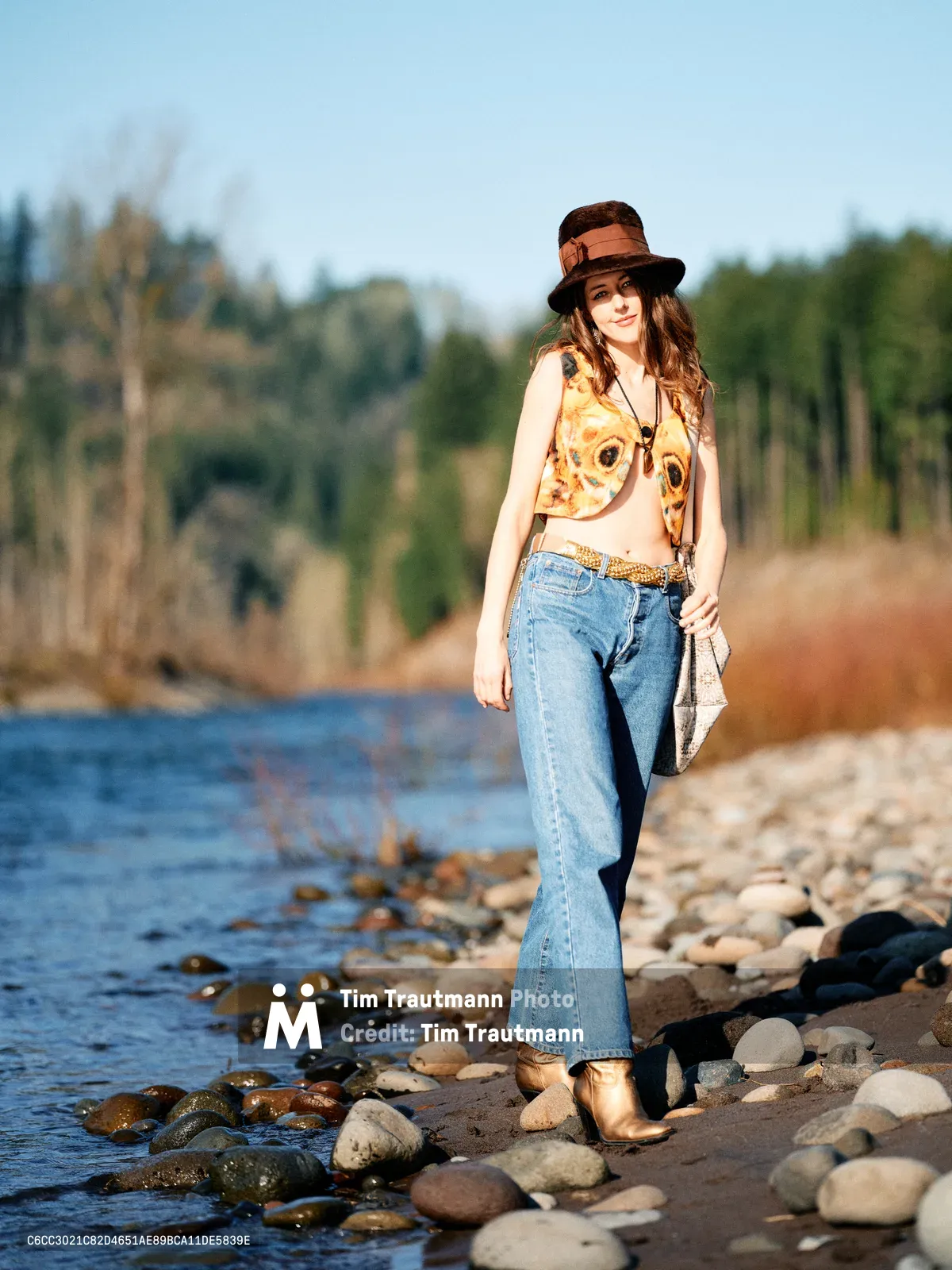 A young woman in bohemian attire stands confidently on a pebbled riverbank along Oregon's Floodplain Trail. Golden hour light illuminates her vintage-inspired ensemble of flared jeans, graphic tank top, and distinctive wide-brimmed hat as she poses beside gently lapping waters. The shallow river meanders through a backdrop of towering evergreens and deciduous trees, creating an intimate wilderness portrait that captures both personal style and natural serenity.