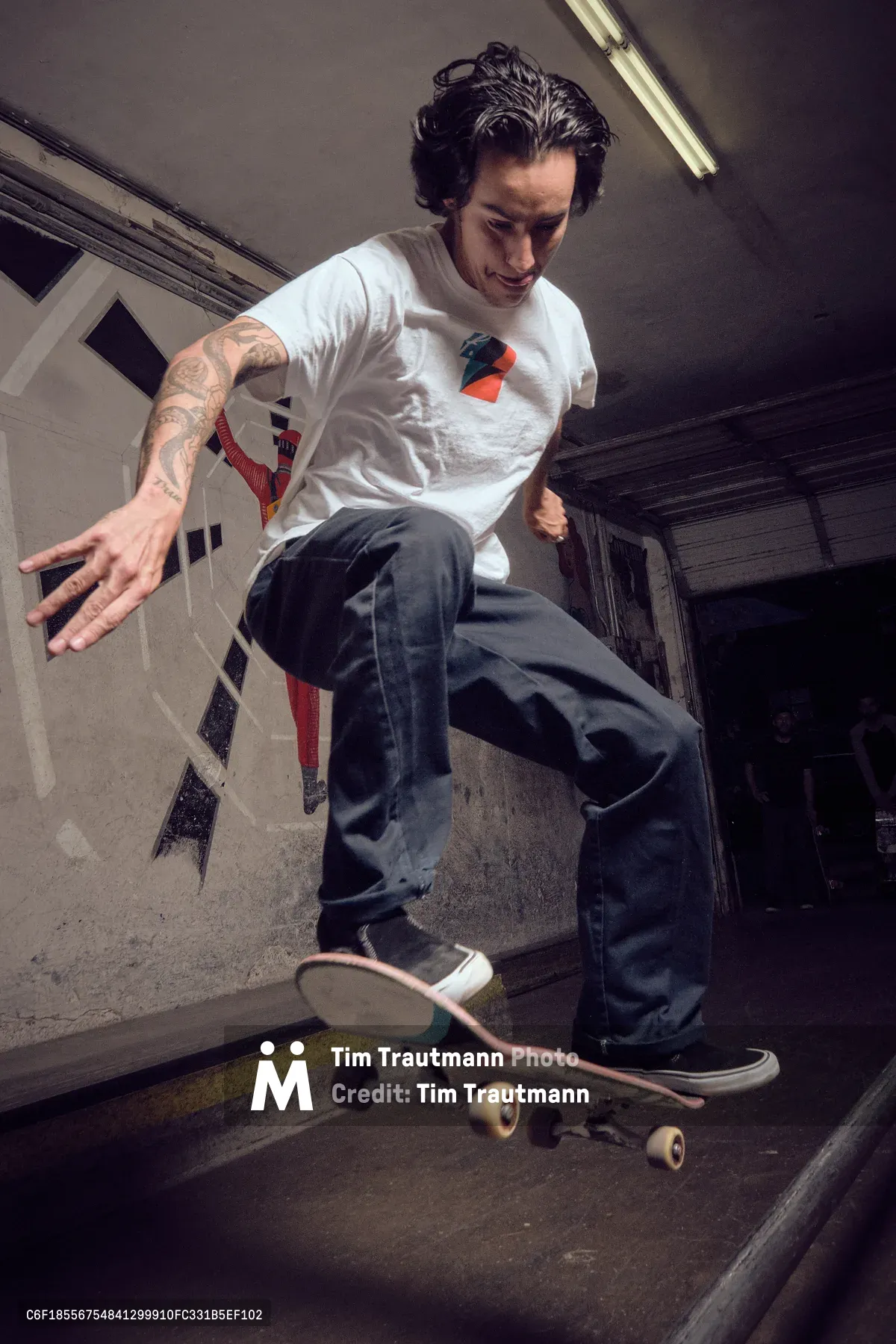 A tattooed skateboarder in a white graphic tee executes a fluid movement within the weathered concrete bowl of Commonwealth Skateboarding in Portland's Buckman neighborhood. Harsh fluorescent lighting cuts through the subterranean atmosphere, casting dramatic shadows across the curved transitions and worn surfaces. The skater's focused expression and extended arm suggest a moment of balance and control as his board glides through the empty pool, embodying the raw authenticity of underground skate culture.