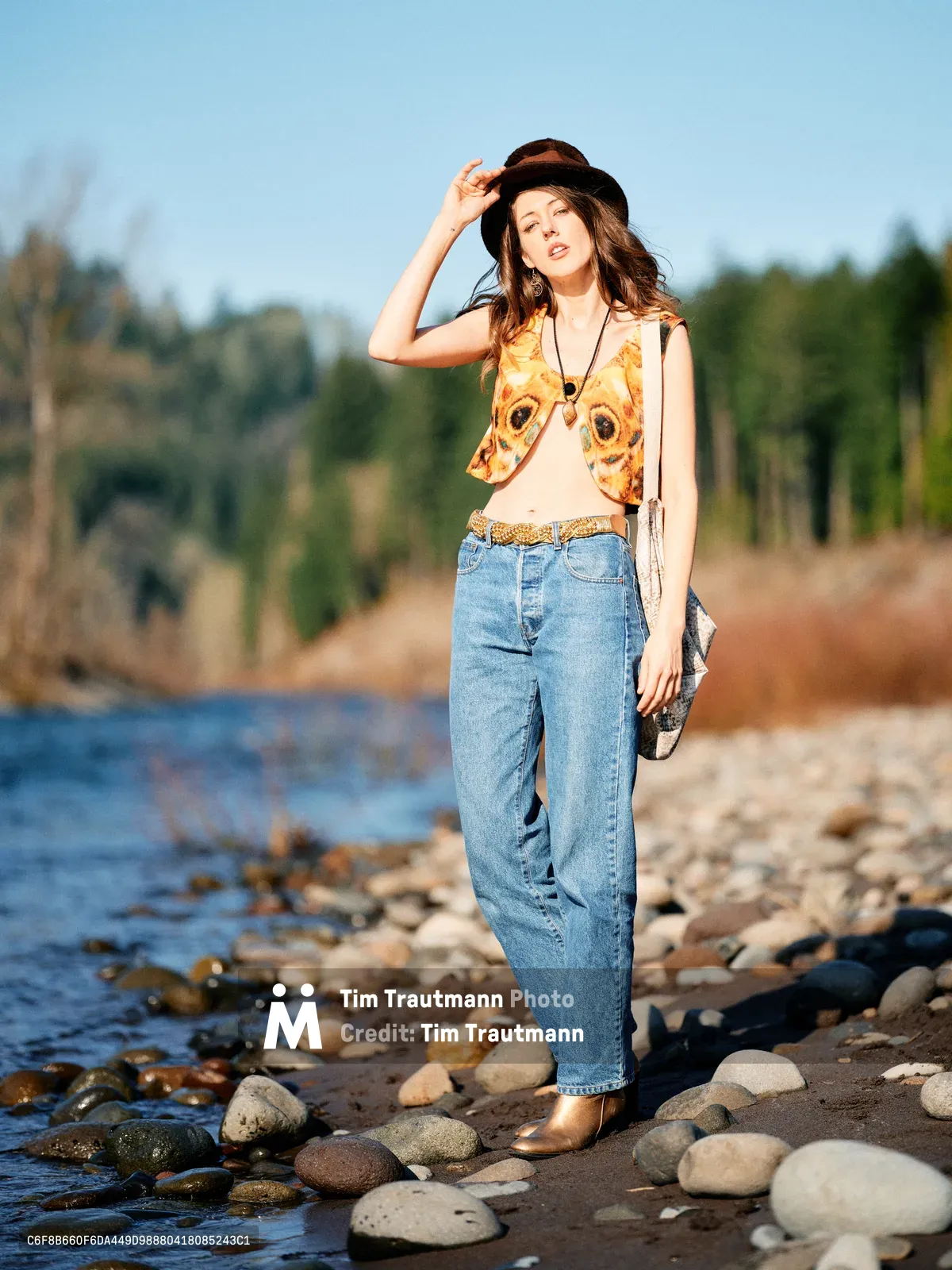 A young woman in bohemian attire poses confidently along the rocky shoreline of the Floodplain Trail in Multnomah County, Oregon. She wears a vibrant butterfly-print vest over a cream top, high-waisted denim, and a dark fedora while touching the brim with one hand. The golden hour light illuminates her flowing hair and creates a warm glow against the serene river backdrop dotted with smooth river stones and forested hills.