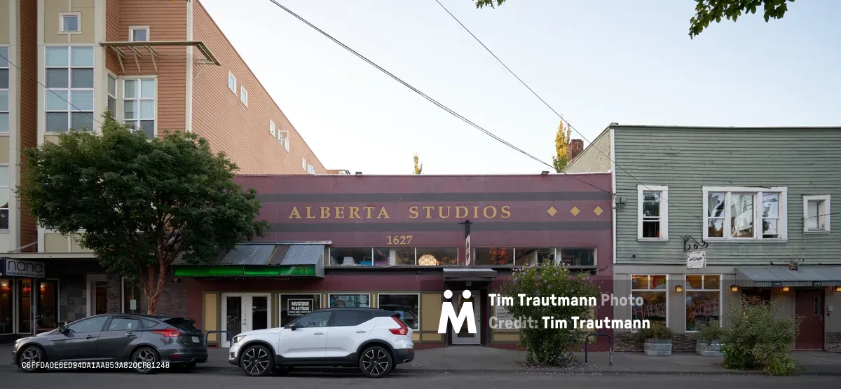 The burgundy facade of Alberta Studios anchors this eclectic stretch of Northeast Alberta Street in Portland's Alberta Arts District. Golden lettering and diamond accents grace the historic building at 1627, while modern vehicles line the street beneath overcast Pacific Northwest skies. The mixed architectural landscape showcases Portland's blend of contemporary development and neighborhood character, with weathered wood siding and large windows creating an inviting creative hub.