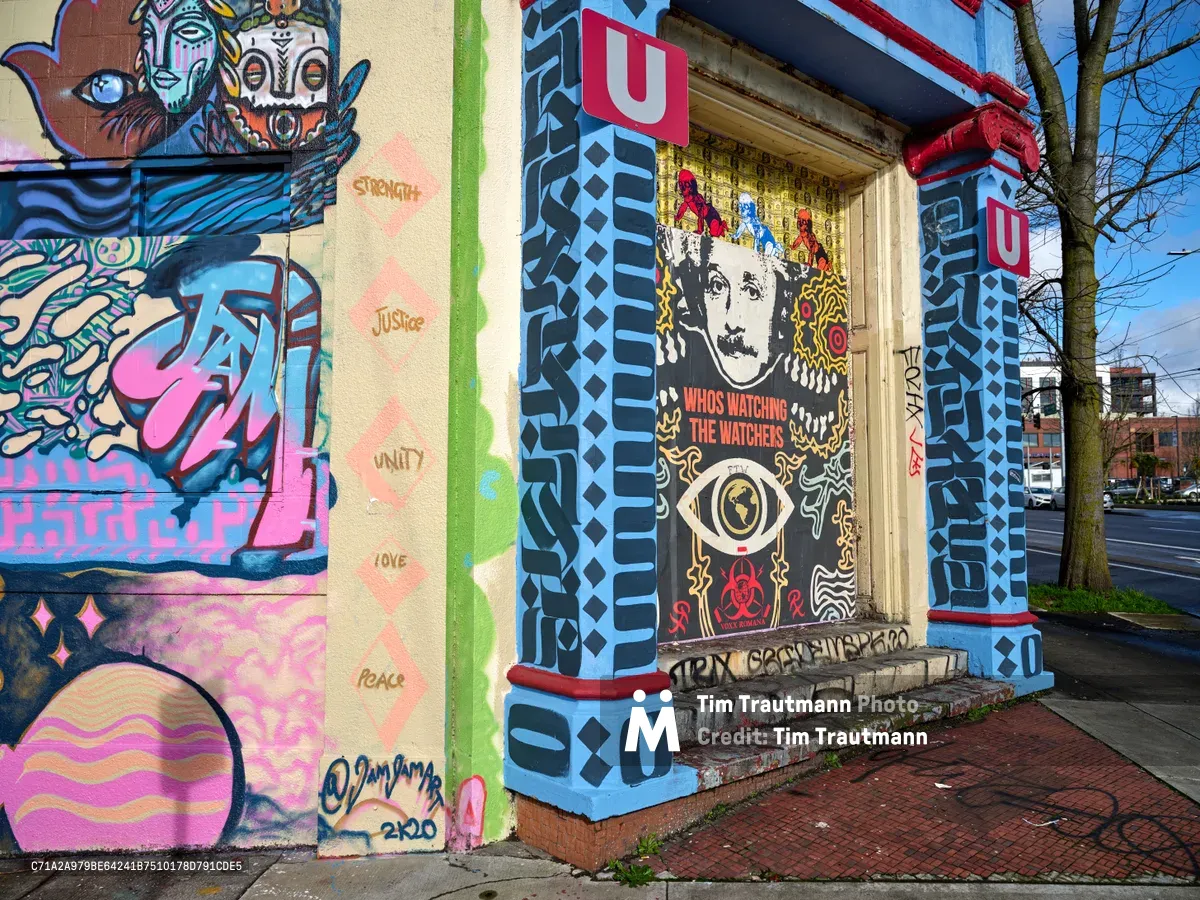 Vibrant street art adorns the columns and walls of the former Salvation Army warehouse in Portland's Central Eastside district, where the Portland Street Art Alliance has commissioned a kaleidoscope of murals. Bold geometric patterns in electric blues frame portraits of Einstein and mythical creatures, while messages of "Strength," "Justice," "Unity," "Love," and "Peace" emerge from pink diamond motifs. The eclectic collision of pop culture iconography, tribal-inspired designs, and surveillance imagery creates a powerful commentary on contemporary urban life against the industrial backdrop of Southeast Ash Street.