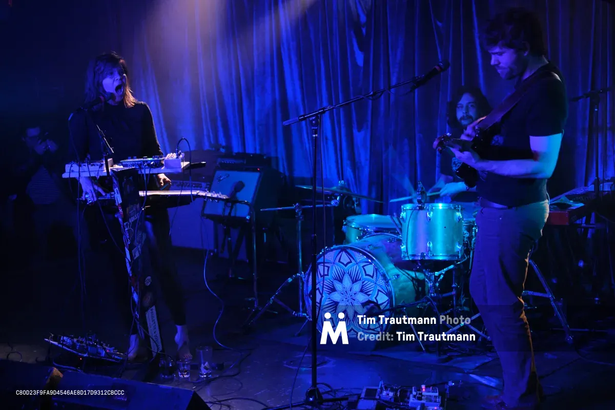The experimental band Sun Blood Stories performs under moody blue stage lighting at Dante's in downtown Portland during Rx Fest. A keyboardist with shoulder-length hair commands the left side of the frame while a guitarist stands prominently on the right, with a drummer visible in the atmospheric shadows between them. The Boise-founded group, known for their opera-like sound and now based in Portland, creates a mesmerizing presence in the intimate venue's draped backdrop and professional lighting that captures the raw energy of their live performance.